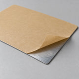 Back of a thin aluminum nameplate with the release liner peeled back to show the adhesive backing